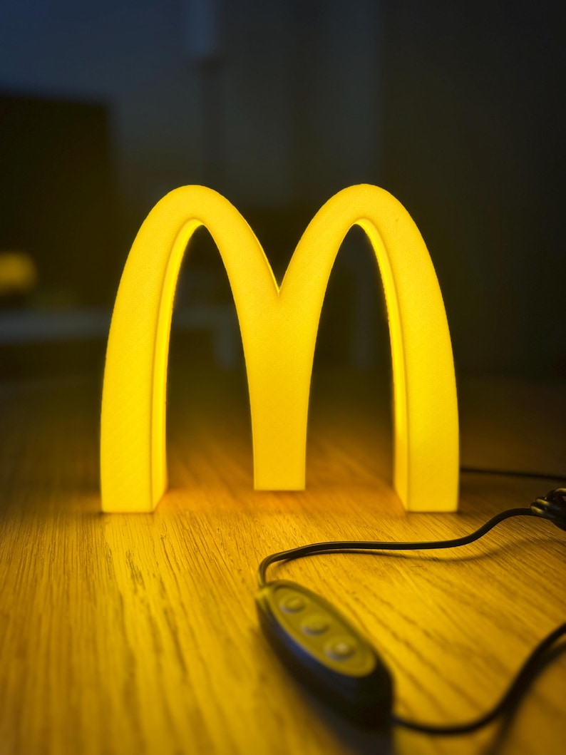 May include: A glowing yellow McDonald's logo, the iconic golden arches, illuminated against a dark background. The light is on a wooden surface, with a black cord and a remote control.