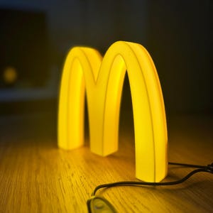 May include: A glowing yellow McDonald's logo in the shape of the letter "M" sits on a wooden surface. The illuminated sign is connected to a black remote control with three buttons. The background is dark.