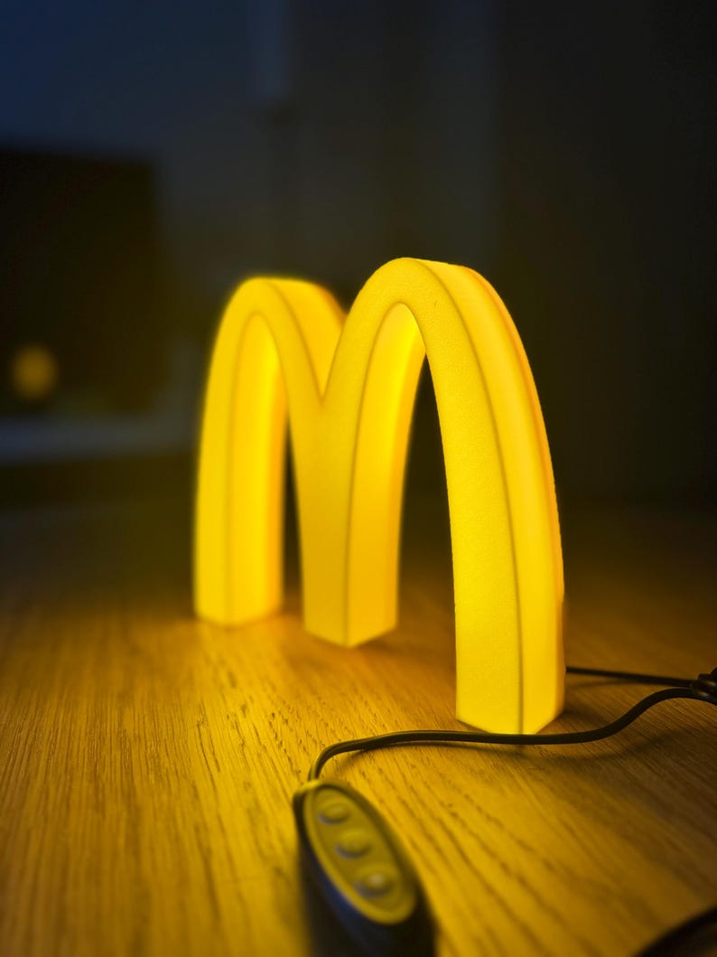 May include: A glowing yellow McDonald's logo in the shape of the letter "M" sits on a wooden surface. The illuminated sign is connected to a black remote control with three buttons. The background is dark.