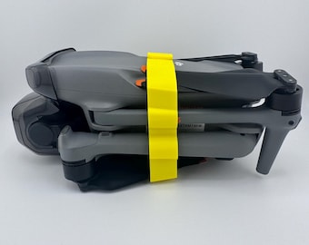 May include: A folded gray drone secured with a bright yellow strap. The drone's propellers are folded, and the strap has orange accents. The drone is on a white background.