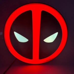 May include: A round, red neon sign featuring the Deadpool logo. The sign has a black background with white, eye-shaped accents. The sign is lit, and the red color is vibrant.