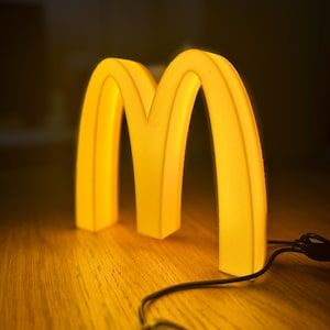May include: A glowing yellow light fixture in the shape of the McDonald's golden arches. The light is on a wooden surface with a black cord and a remote control.