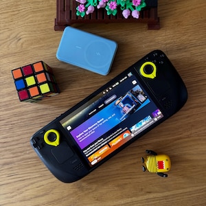 May include: A black handheld gaming device with yellow accents, displaying a game interface. A Rubik's Cube, a light blue wireless charger, and a yellow toy are also present. A small brown decorative planter with pink and purple flowers is in the background.
