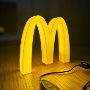 May include: A glowing yellow McDonald's logo in the shape of two golden arches. The illuminated sign is on a wooden surface, with a black cord and remote control visible. The background is dark.