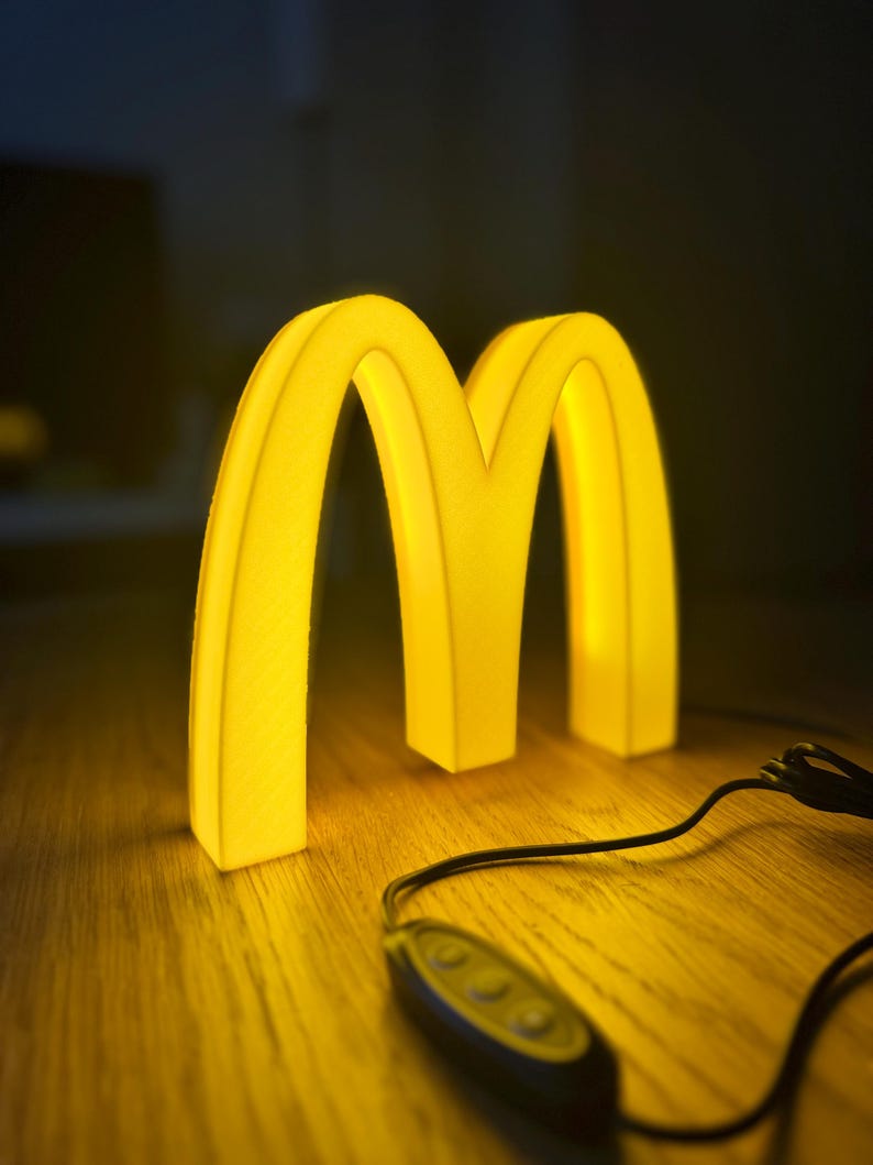 May include: A glowing yellow McDonald's logo in the shape of two golden arches. The illuminated sign is on a wooden surface, with a black cord and remote control visible. The background is dark.