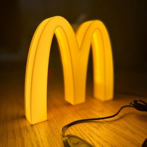 May include: A glowing yellow McDonald's logo in the shape of two arches, illuminated against a dark background. The light is warm and inviting, with a control device in the foreground. The arches are on a wooden surface.