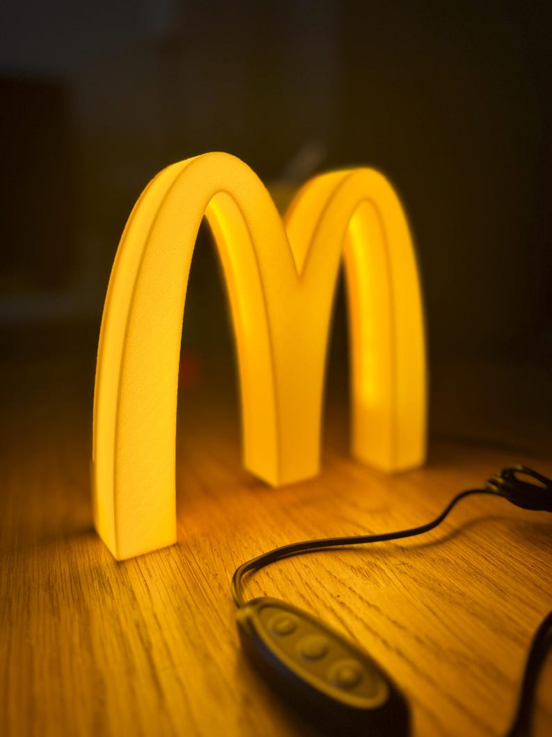 May include: A glowing yellow McDonald's logo in the shape of two arches, illuminated against a dark background. The light is warm and inviting, with a control device in the foreground. The arches are on a wooden surface.