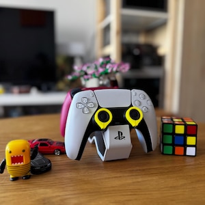 May include: A white and black PlayStation 5 controller with yellow accents, resting on a white charging stand. A Rubik's Cube, toy cars, and a yellow cartoon figure are also on the wooden surface.