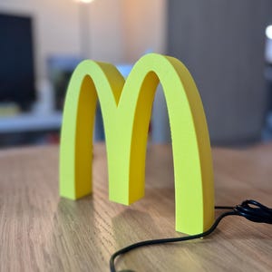 May include: A bright yellow, three-dimensional sculpture of the McDonald's golden arches logo. The sculpture is on a wooden surface, with a black cord and a remote control in the foreground. The background is blurred.