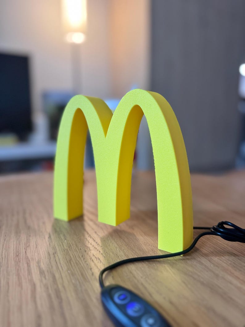 May include: A bright yellow, three-dimensional sculpture of the McDonald's golden arches logo. The sculpture is on a wooden surface, with a black cord and a remote control in the foreground. The background is blurred.