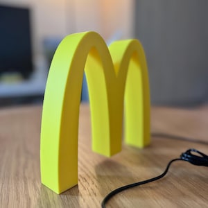 May include: A bright yellow, three-dimensional McDonald's logo, the iconic golden arches, stands on a wooden surface. A black cable with a control panel is in the foreground. The background is blurred, with a glimpse of a monitor.