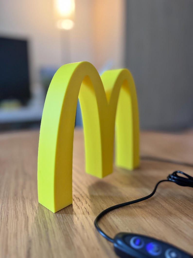 May include: A bright yellow, three-dimensional McDonald's logo, the iconic golden arches, stands on a wooden surface. A black cable with a control panel is in the foreground. The background is blurred, with a glimpse of a monitor.
