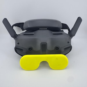 May include: A black VR headset with two antennas and a yellow lens cover. The headset has a head strap and is designed for virtual reality experiences. The yellow lens cover is in the shape of glasses.