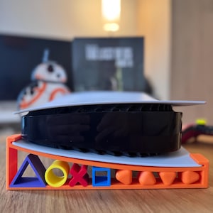 Może przedstawiać: Czarno-biała konsola do gier spoczywa na kolorowej podstawce. Podstawka jest pomarańczowa z niebieskimi, żółtymi, czerwonymi i pomarańczowymi kształtami geometrycznymi. Konsola ma białą górę i czarny środek.