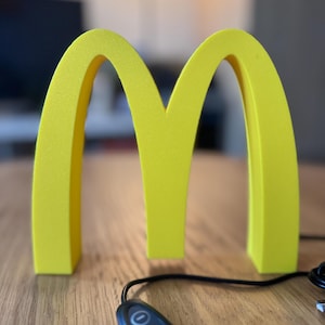 May include: A bright yellow, three-dimensional McDonald's logo, the iconic golden arches, stands on a wooden surface. A black remote control with three buttons and a cord is in the foreground. The background is blurred.