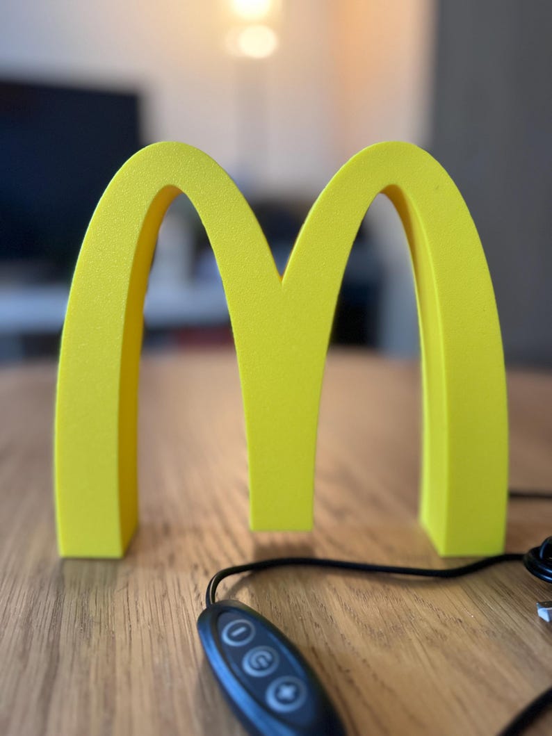 May include: A bright yellow, three-dimensional McDonald's logo, the iconic golden arches, stands on a wooden surface. A black remote control with three buttons and a cord is in the foreground. The background is blurred.