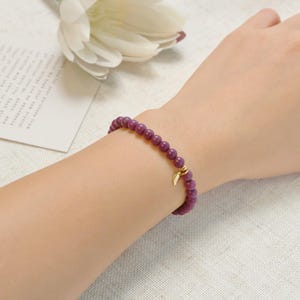 May include: A beaded bracelet with small, round, purple beads. A gold-colored charm in the shape of a leaf hangs from the bracelet. The bracelet is worn on a wrist.