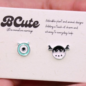 May include: A pair of ultra-miniature earrings on a white card. One earring is a teal-coloured eye design, and the other is a black and white face design. The card has the text "BCute" and "Adorable plant and animal designs".