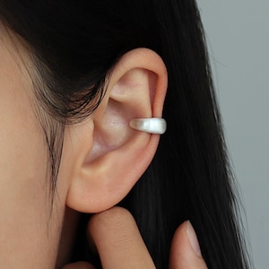 Peut inclure: Un ear cuff argenté est porté sur l'oreille. Le ear cuff a un design simple et incurvé. L'arrière-plan est de couleur grise unie. Les cheveux de la personne sont noirs.
