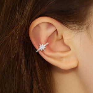 May include: A silver ear cuff with a starburst design, set with small, sparkling stones. The cuff curves around the ear cartilage, with more stones along the band. The jewellery is shown on a person's ear.