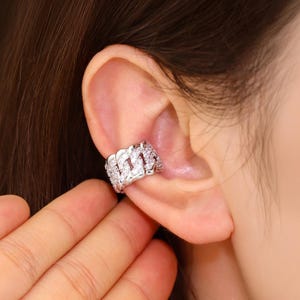 Può includere: Un ear cuff argentato con un design a catena, incrostato con piccole pietre scintillanti. Il gioiello è indossato sull'orecchio, mostrando i suoi dettagli intricati e la superficie riflettente. Il gioiello aggiunge un tocco di eleganza.