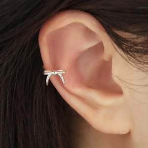 Op de afbeelding: Een zilveren oormanchet in de vorm van een strik wordt op het oor gedragen. De sieraden zijn gemaakt van een glanzend, metalen materiaal. De oormanchet is een delicaat accessoire.