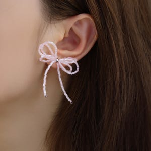 May include: A close-up of a pink beaded bow earring. The earring is attached to an ear. The bow is made of small, light pink beads and has two long, dangling strands. The earring has a silver-coloured post.