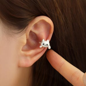 May include: A silver cat ear cuff earring. The cat design features small, round eyes and a tiny nose. The earring is worn on the ear.