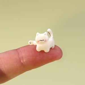 Puede incluir: Una pequeña figura de gato blanco con una pata levantada y una cola curvada. El gato tiene mejillas rosadas y puntos negros para los ojos y la nariz. La figura está sobre un dedo contra un fondo verde claro.