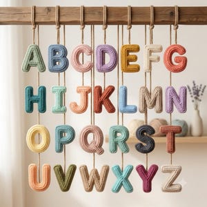 Puede incluir: Un conjunto de veintiséis letras de ganchillo de la A a la Z, cada una elaborada en un color de hilo pastel o vibrante diferente. Las letras están suspendidas por cordel marrón fino de una viga de madera horizontal, creando un alfabeto colgante.