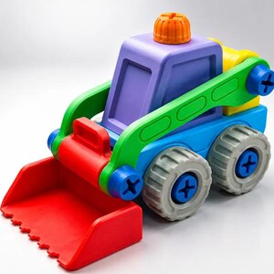 Op de afbeelding: Een kleurrijk plastic speelgoedbouwvoertuig met een rode voorbak, een paarse cabine met een oranje knop aan de bovenkant, groene steunarmen en een blauw chassis. Het voertuig heeft vier grijze wielen met blauwe kruiskopschroeven.