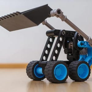 Könnte beinhalten: Ein Baufahrzeug-Spielzeug aus schwarzen, blauen und grauen Kunststoff-Bausteinen. Das Modell verfügt über eine große schwarze Frontschaufel, einen mechanischen Arm und vier Räder mit schwarzen Profilreifen und hellblauen Radnaben.