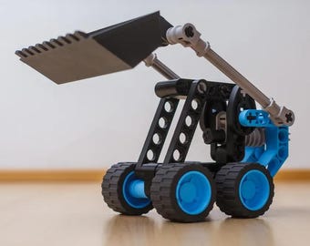 May include: A construction vehicle toy built from black, blue, and gray plastic building blocks. The model features a large black front bucket, a mechanical arm assembly, and four wheels with black treads and bright blue inner hubs.