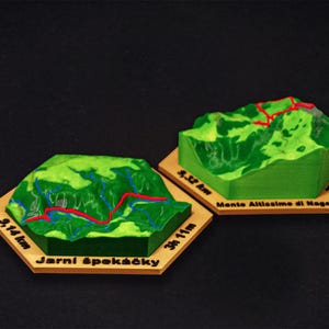 May include: Two miniature green mountain models with red and blue trail markings sit on hexagonal wooden bases. One base reads "9.14 km" and "3h 11m". The other reads "9.52 km" and "3h 57m".