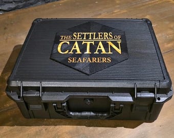 May include: A black rectangular hard-shell carrying case with a ribbed texture and a central hexagonal emblem. The emblem features the gold text The Settlers of Catan Seafarers. The case includes a sturdy handle and secure locking latches.