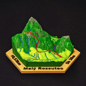 May include: A 3D-printed topographical model of a mountain, featuring green terrain with a red hiking trail. Blue lines represent streams. The base is tan with the text "Malý Rozsutec" and measurements of 10.53km and 5h 39m.