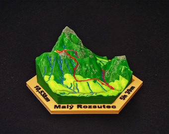 May include: A 3D-printed topographical model of a mountain, featuring green terrain with a red hiking trail. Blue lines represent streams. The base is tan with the text "Malý Rozsutec" and measurements of 10.53km and 5h 39m.