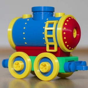 Peut inclure: Une locomotive de train jouet construite en briques de plastique. Le corps cylindrique principal est bleu avec des bordures jaunes et une face avant rouge. Elle comporte une échelle jaune sur le côté, une cheminée bleue et quatre roues jaunes.