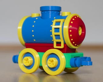 May include: A toy train engine constructed from plastic building blocks. The main cylindrical body is blue with yellow trim and a red front. It features a yellow ladder on the side, a blue chimney on top, and four yellow wheels attached to a green base.
