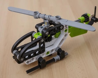 May include: A construction toy helicopter built from interlocking plastic beams and connectors. The model features a white, black, and lime green color scheme with a gray main rotor blade assembly and a small black tail rotor at the rear.
