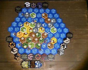 May include: A Catan Seafarers board game setup featuring hexagonal tiles in blue, yellow, green, and brown. The board includes numbered tokens, small wooden game pieces, and various resource cards arranged in a large hexagonal pattern with Catan branding.