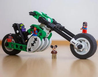 May include: A custom-built motorcycle constructed from green, black, and white plastic building blocks. The vehicle features a long front fork, large black tires, and several small character figurines positioned on the frame and front wheel.