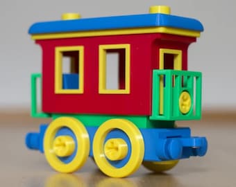 May include: A toy train car constructed from plastic building blocks. The main body is red with yellow window frames, a blue roof, and a green balcony. The base is green with blue accents and four yellow wheels with blue centers.