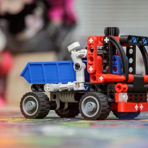 Op de afbeelding: Een speelgoed kiepwagen gebouwd van in elkaar grijpende kunststof onderdelen. Het voertuig heeft een felblauwe laadbak, een rood en zwart cabineframe en vier zwarte wielen met grijze centra. Een witte mechanische arm is aan de zijkant bevestigd.