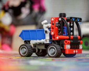 May include: A construction toy dump truck built from interlocking plastic pieces. The vehicle features a bright blue cargo bed, a red and black cab frame, and four black wheels with grey centers. A white mechanical arm is attached to the side.