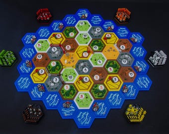 May include: A board game featuring a hexagonal grid of terrain tiles including green forests, yellow wheat fields, brown hills, and gray mountains, surrounded by blue water tiles. Small plastic game pieces in red, orange, green, white, black, and yellow are arranged around the board.