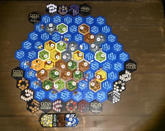 May include: A Catan Seafarers board game setup featuring hexagonal tiles in blue, yellow, green, and brown. The board includes numbered tokens, small wooden game pieces, and various resource cards arranged in a large hexagonal pattern with Catan branding.