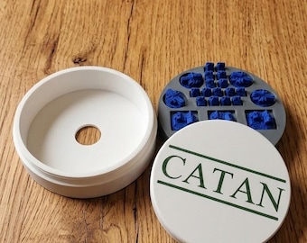 May include: A white container with a central hole, a gray tray with blue game pieces, and a white disc with "CATAN" in green text. The items are on a wooden surface, likely for the board game Catan.