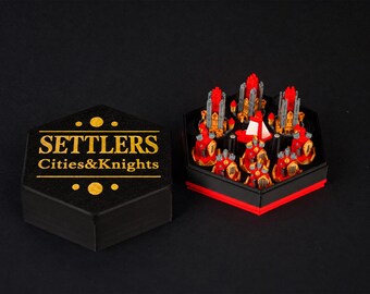 May include: A hexagonal black storage box with the gold text SETTLERS Cities&Knights on the lid, placed next to an open hexagonal container filled with miniature red and gold plastic game pieces including towers and a small white ship.