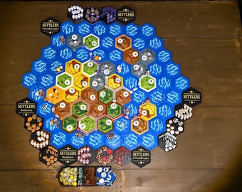 May include: A board game setup featuring a hexagonal map with blue water tiles surrounding land tiles in green, yellow, and brown. The board includes numbered tokens, small game pieces, and black hexagonal border tiles labeled Settlers.